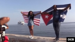 Dos cubanos, con las banderas de Cuba y Estados Unidos, esperan la llegada del Adonia en el Malecón habanero.